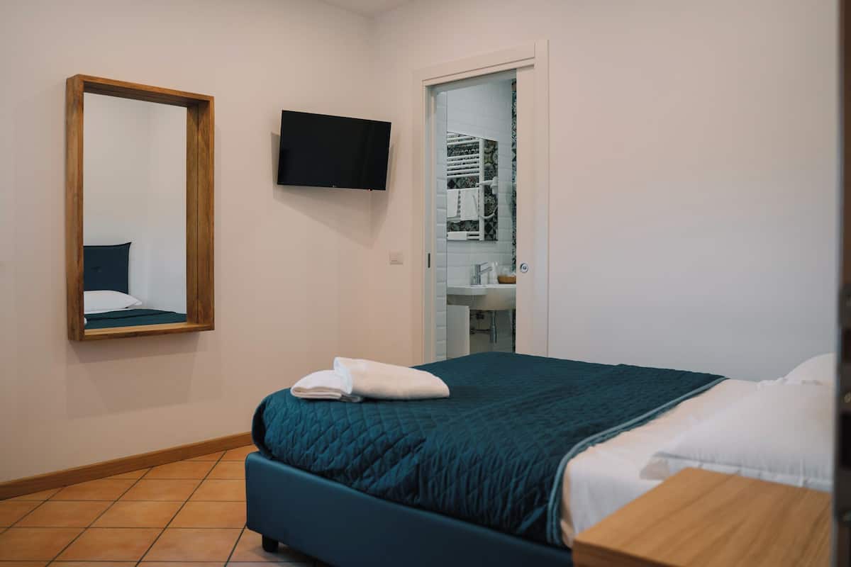 Une chambre avec un lit, un téléviseur fixé au mur, une salle de bain visible à travers une porte ouverte et un sol carrelé.
