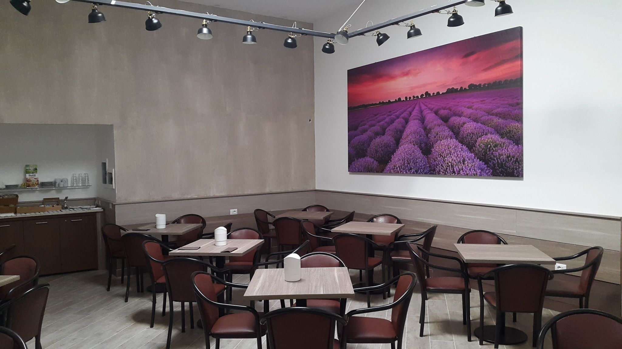 Un espace repas avec des tables et des chaises, un grand tableau représentant des champs de lavande, et un bar avec des bouteilles et des verres.