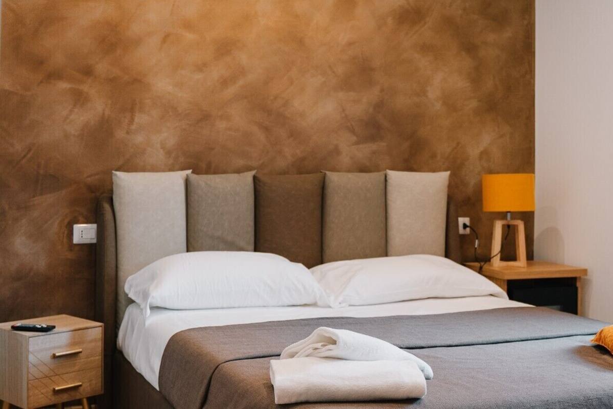 Un lit bien fait, avec du linge de lit blanc, une table de chevet en bois surmontée d’une lampe, et un mur orné d’un motif texturé.
