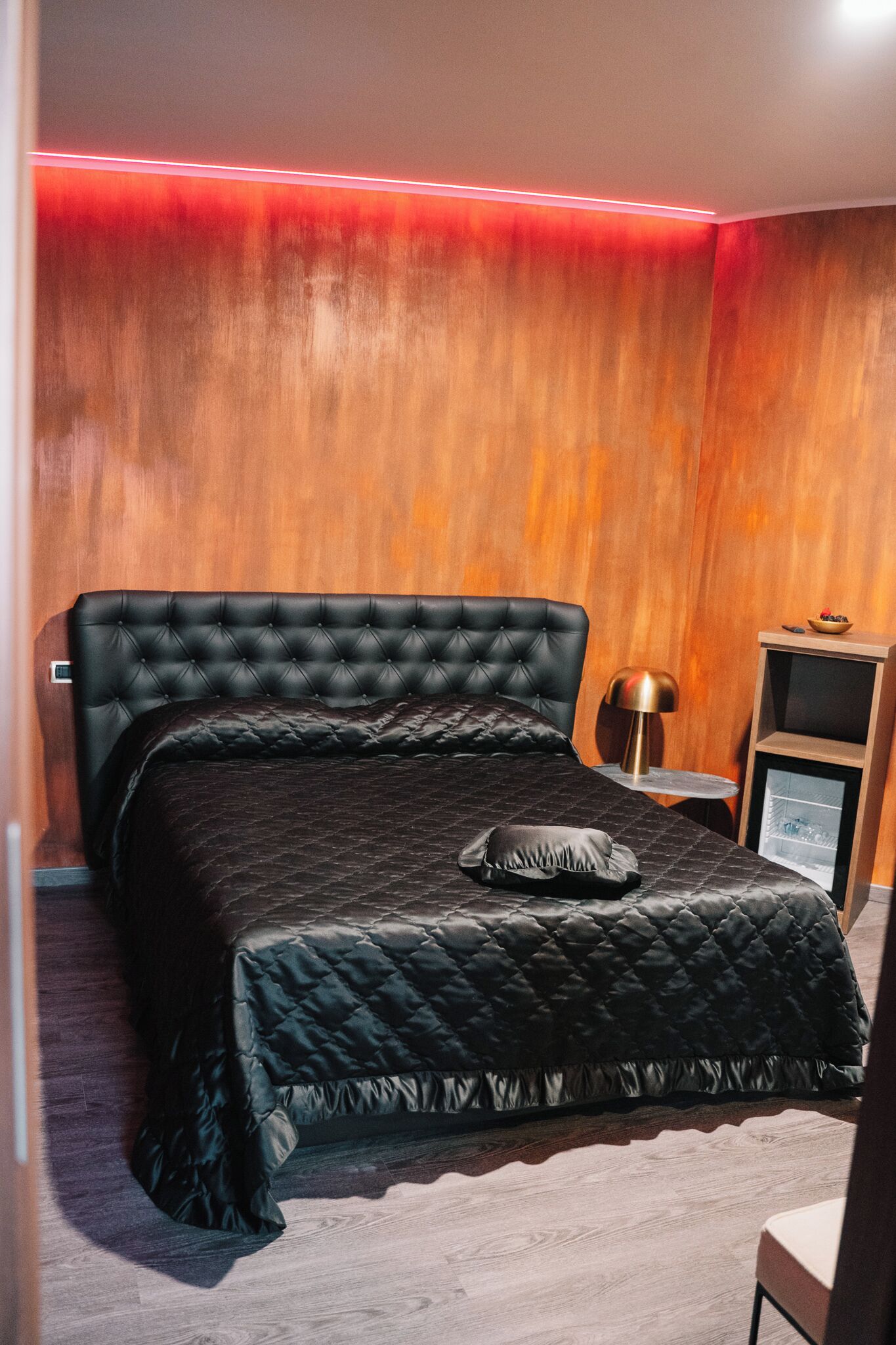 Une chambre à coucher avec un lit noir matelassé, une petite table de chevet avec une lampe et un mur en bois agrémenté d’une bande lumineuse rouge.