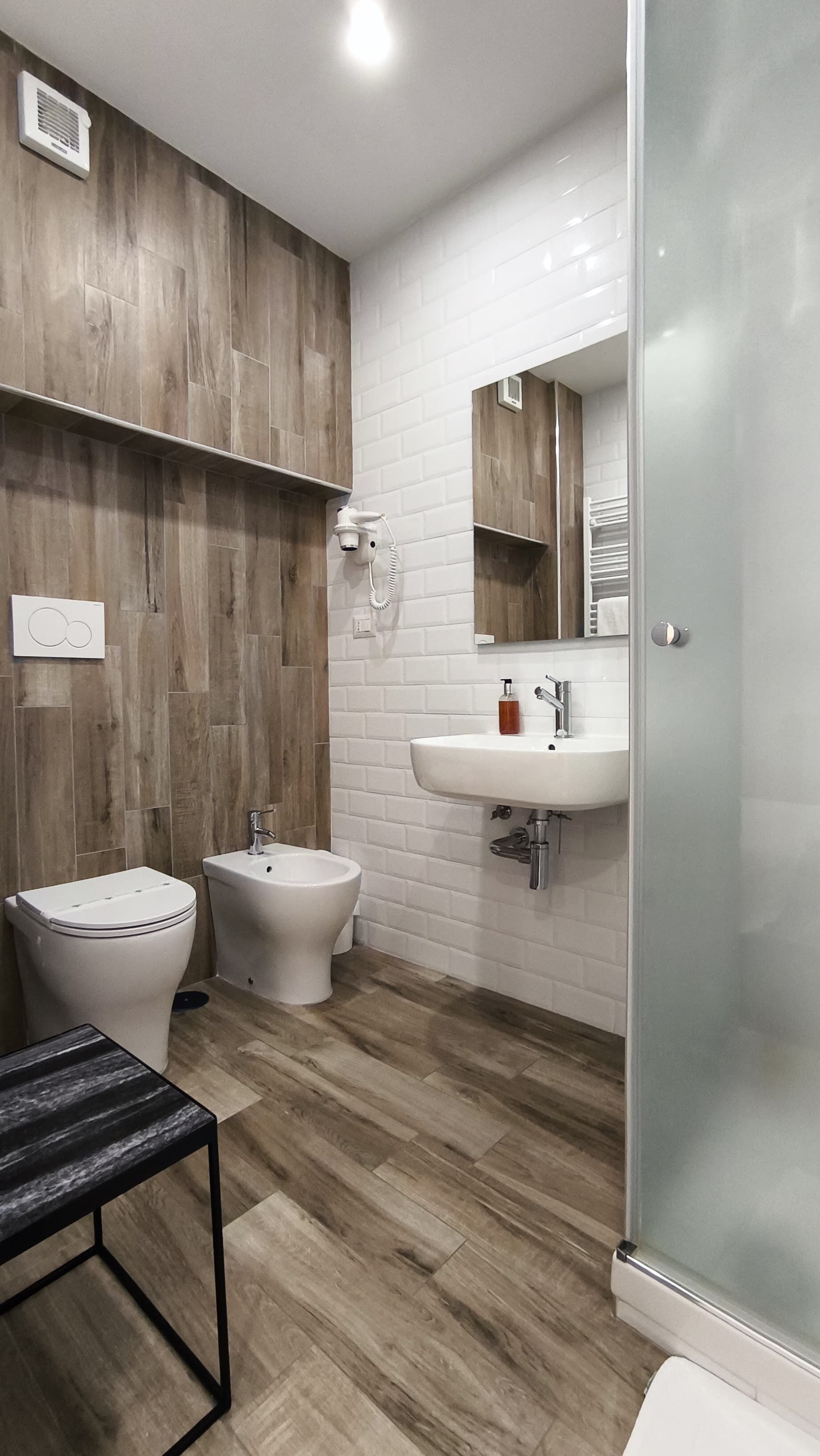 A modern bathroom with a wooden wall panel, white tiles, a sink, a bidet, and a glass shower enclosure.