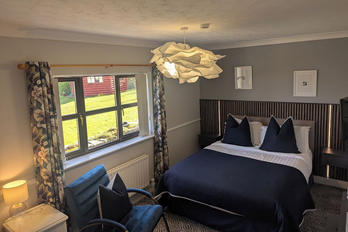 A bedroom with a bed, a chair, a nightstand, a lamp, a window with curtains, and a ceiling light fixture.