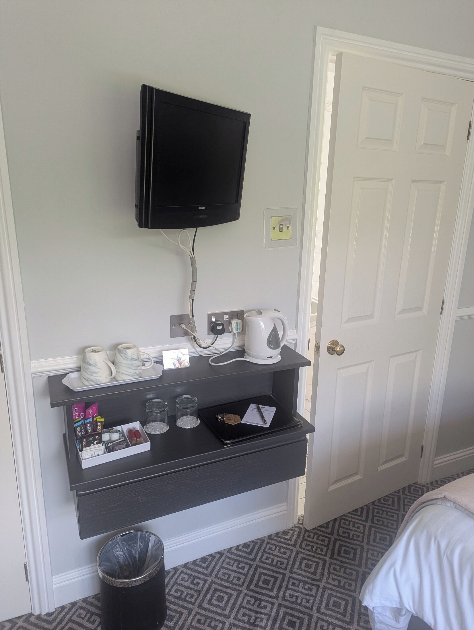 A hotel room with a flat-screen TV mounted on the wall, a coffee station with mugs and a coffee maker, a small shelf with toiletries, and a door with a patterned carpet.
