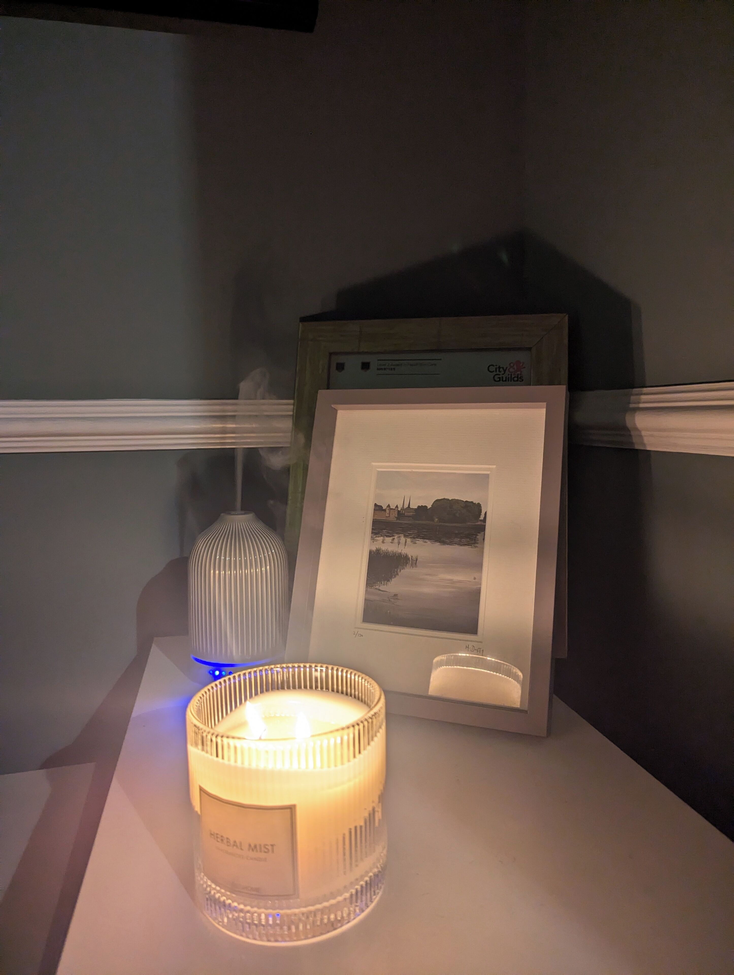 Una vela con etiqueta, un cuadro en marco y un jarrón decorativo sobre una mesa.