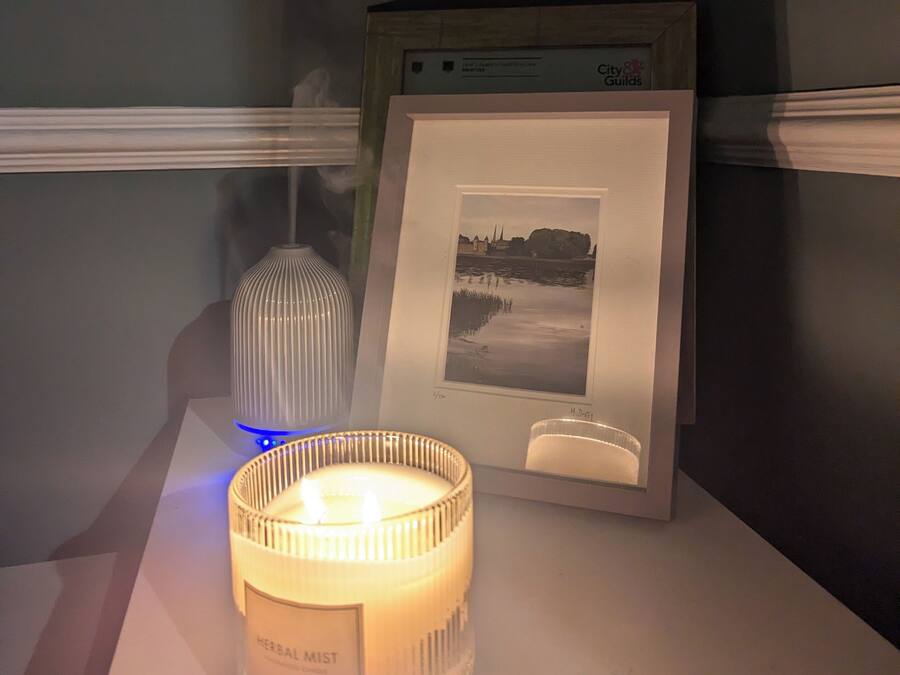 Una vela con etiqueta, un cuadro en marco y un jarrón decorativo sobre una mesa.