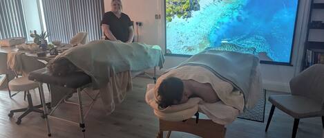 Ein Raum mit zwei Massagetischen, einem großen Bildschirm, der eine Strandansicht zeigt, und einer Person, die am Fenster steht.