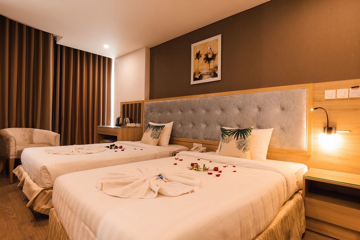 A hotel room with two beds, a large headboard, a nightstand, a desk, and a wall-mounted lamp.