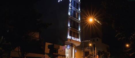 Un hotel de varios pisos con señalización iluminada, entrada visible y vista a la calle con edificios circundantes.