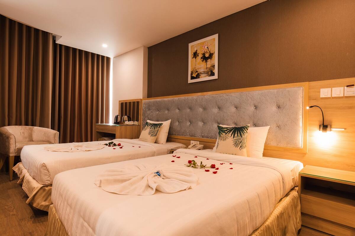 A hotel room with two beds, a large headboard, a bedside table, a chair, and a wall-mounted artwork.