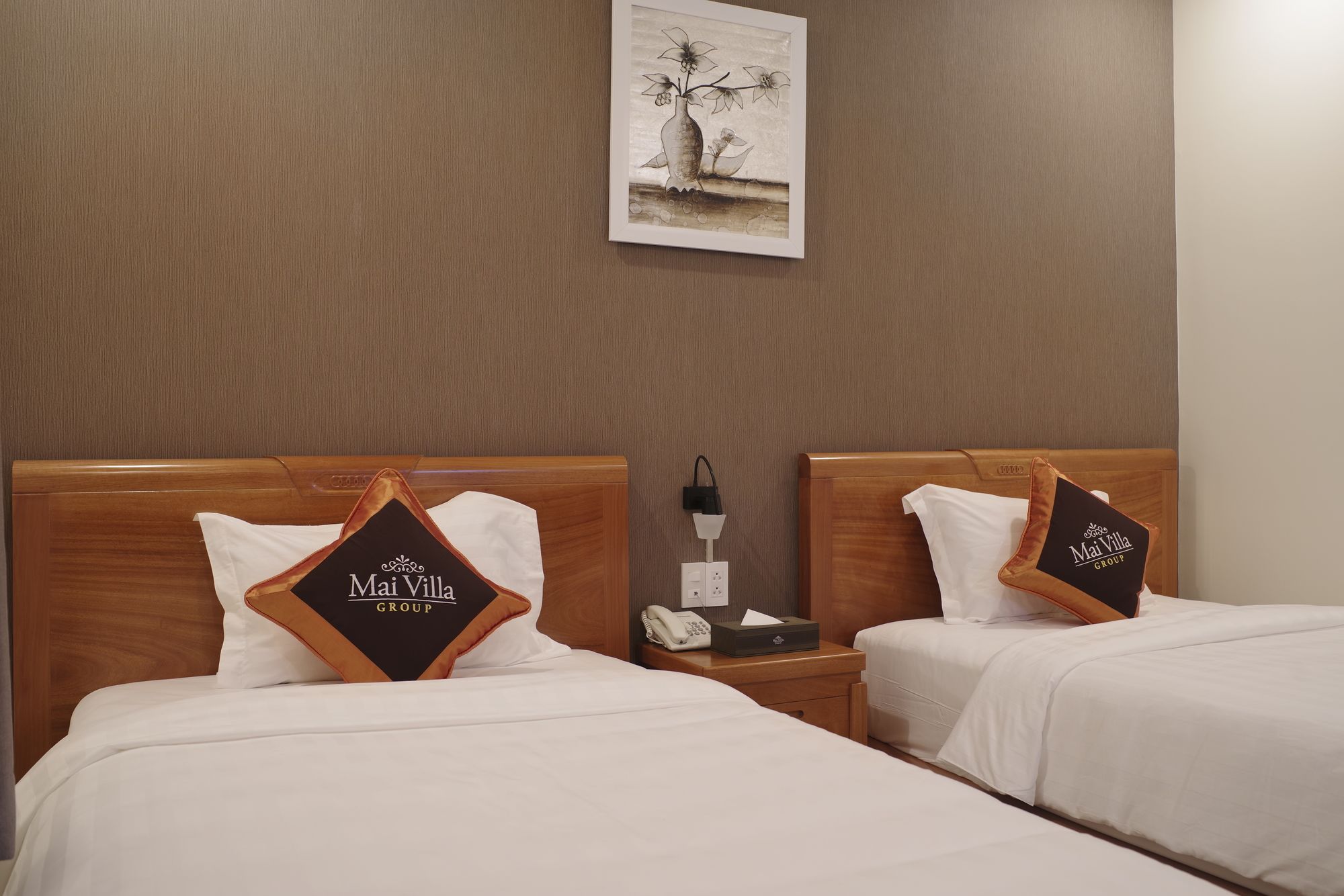 A hotel room with two beds, each with a pillow featuring the hotel's logo, a bedside table with a lamp, and a wall-mounted artwork.