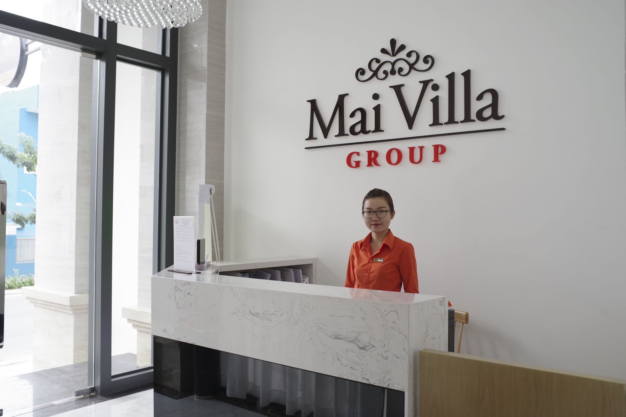 Una mesa de recepción con una placa con el nombre Mai Villa Group, un interior moderno con encimera de mármol y un amplio ventanal que permite la entrada de luz natural.