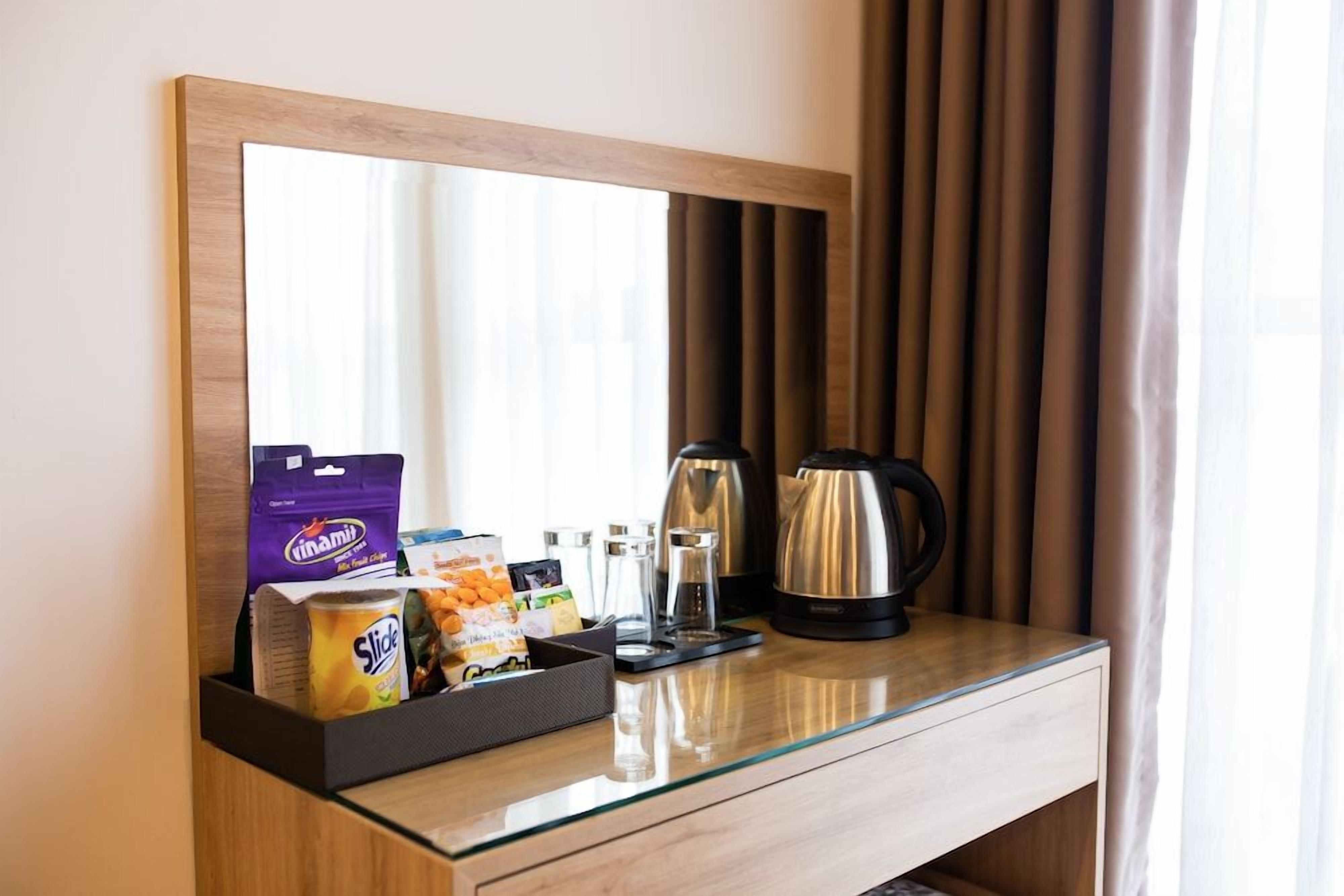 Habitación de hotel con un escritorio de madera, un espejo, una cafetera, un hervidor de agua y una bandeja de snacks con aperitivos y una bebida.