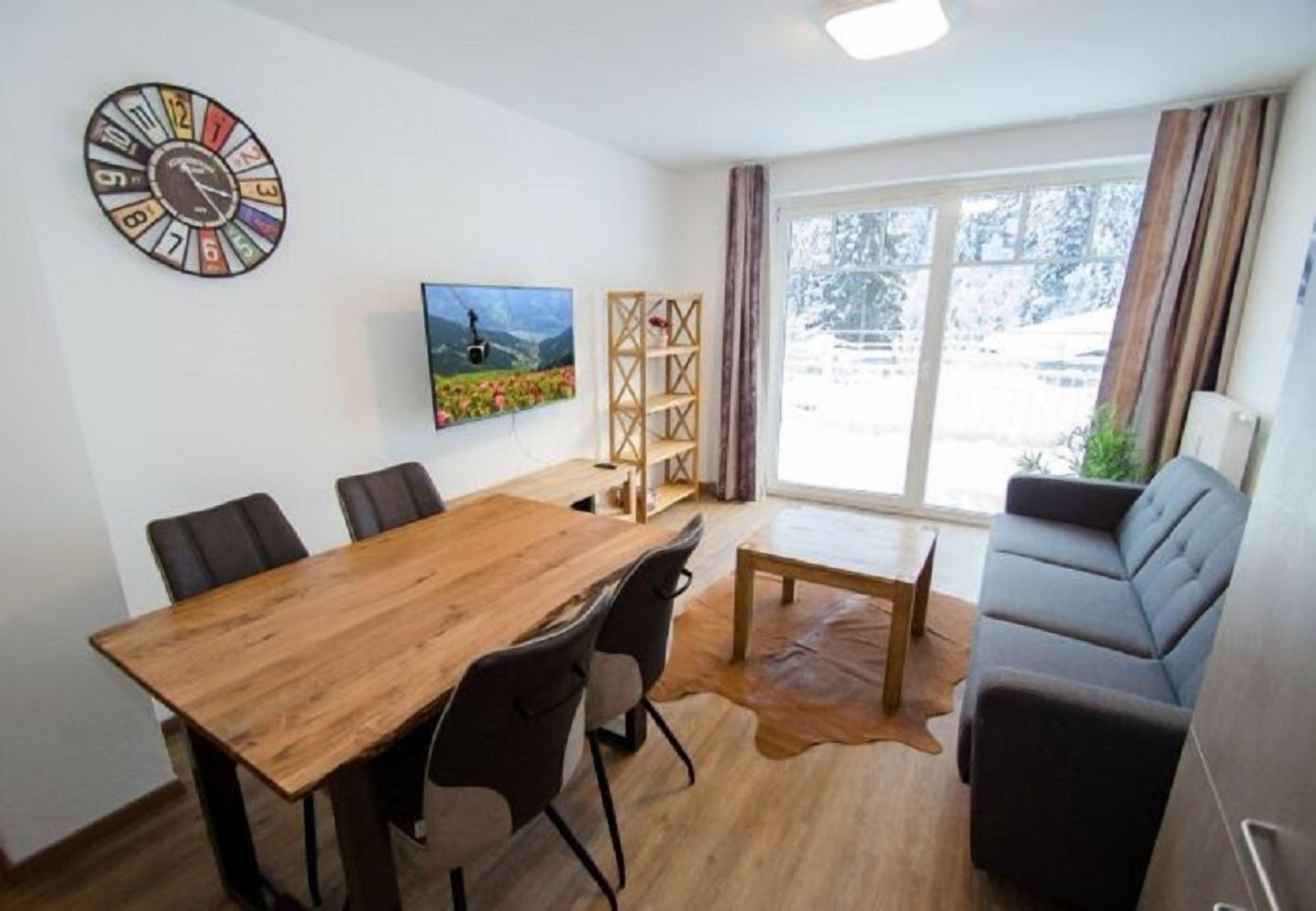 A modern living room with a wooden dining table, leather sofa, and a large window with a view of snow-covered trees.