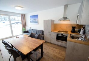 Une cuisine moderne avec une table et des chaises en bois, un four encastré et une vue sur l’extérieur grâce à de grandes fenêtres.