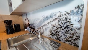 Cozinha com pia de aço inoxidável, máquinas de café pretas e painel decorado com tema de montanha.