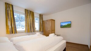 Quarto de hotel com duas camas, um guarda-roupa de madeira, uma janela com cortinas e um quadro de paisagem na parede.