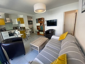 Uma sala de estar moderna com sofá, mesa de centro, TV e um espaço de refeições com cadeiras, além de uma cozinha ao fundo.