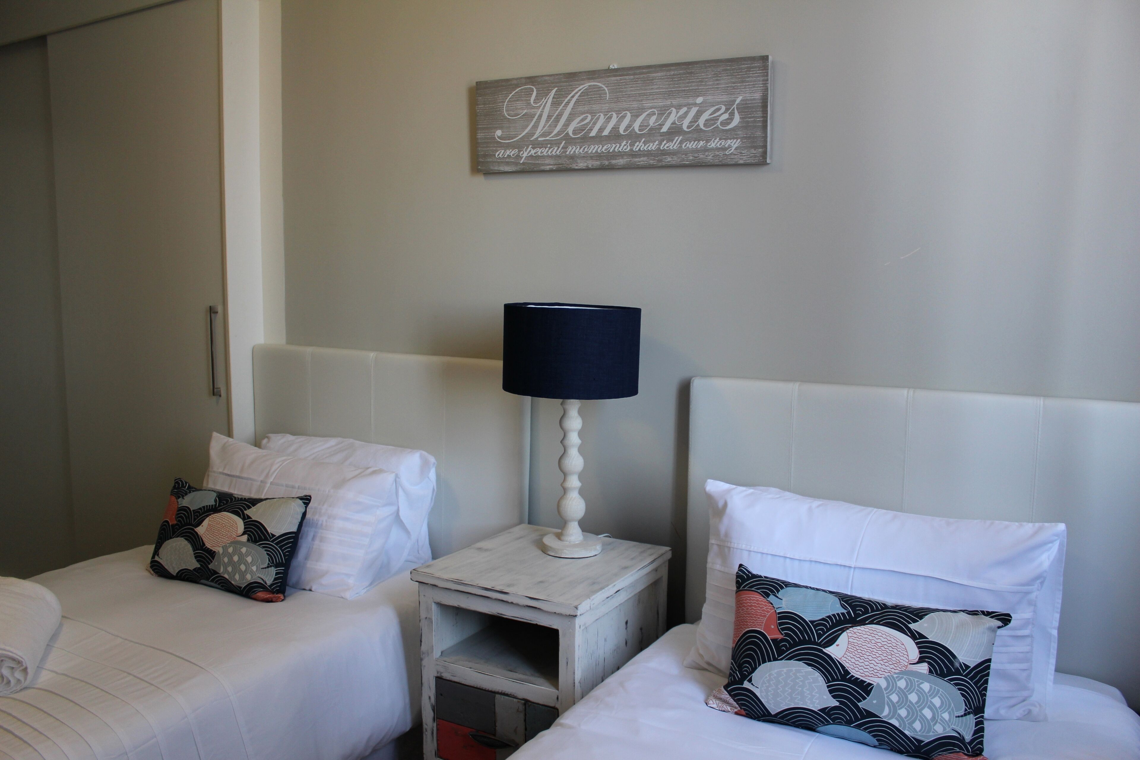 A hotel room with two beds, each with a pillow and a bedside table with a lamp. There is a wall sign above the beds.