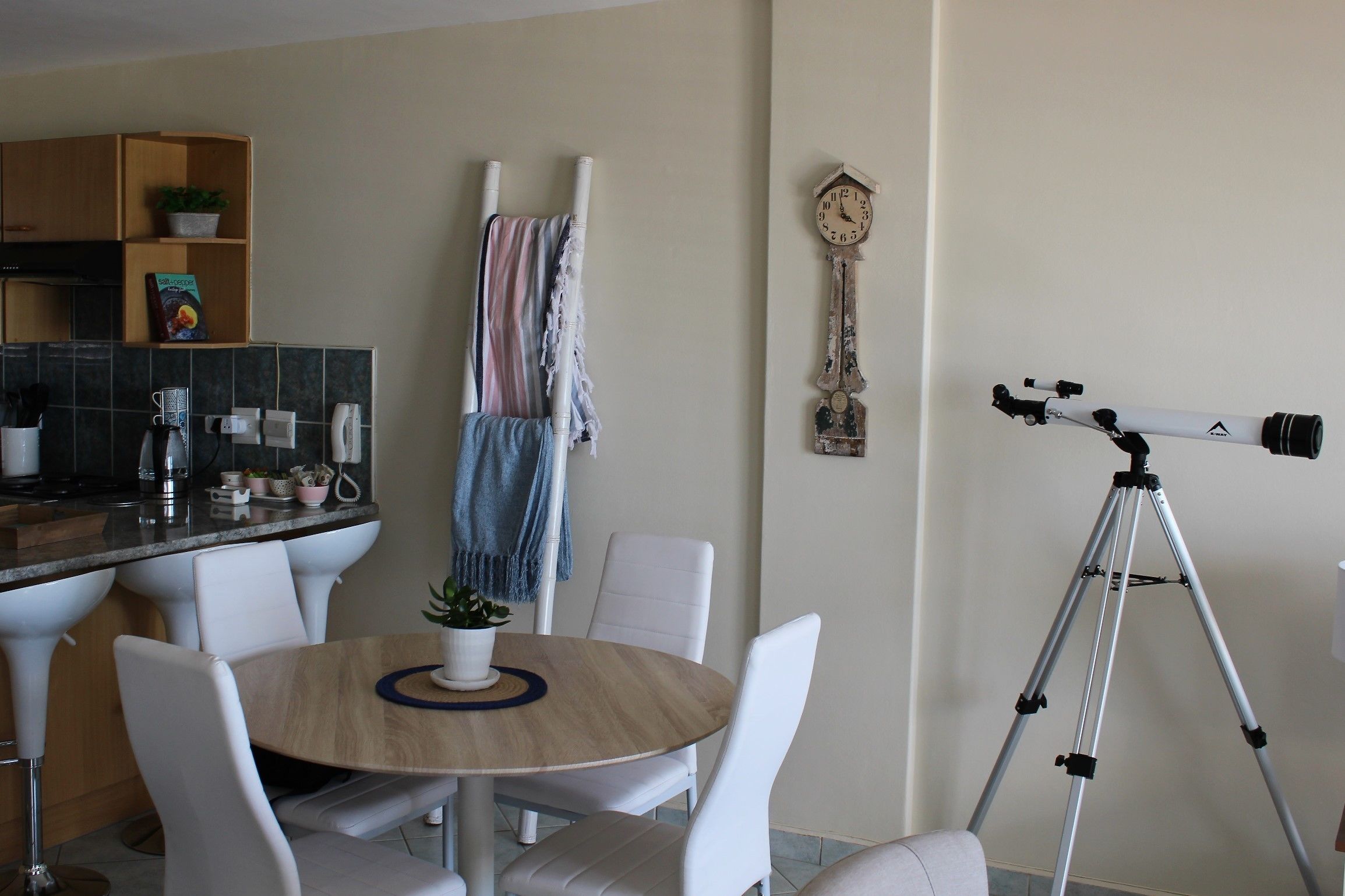Un comedor con una mesa redonda, sillas blancas, un reloj y un telescopio sobre un trípode.