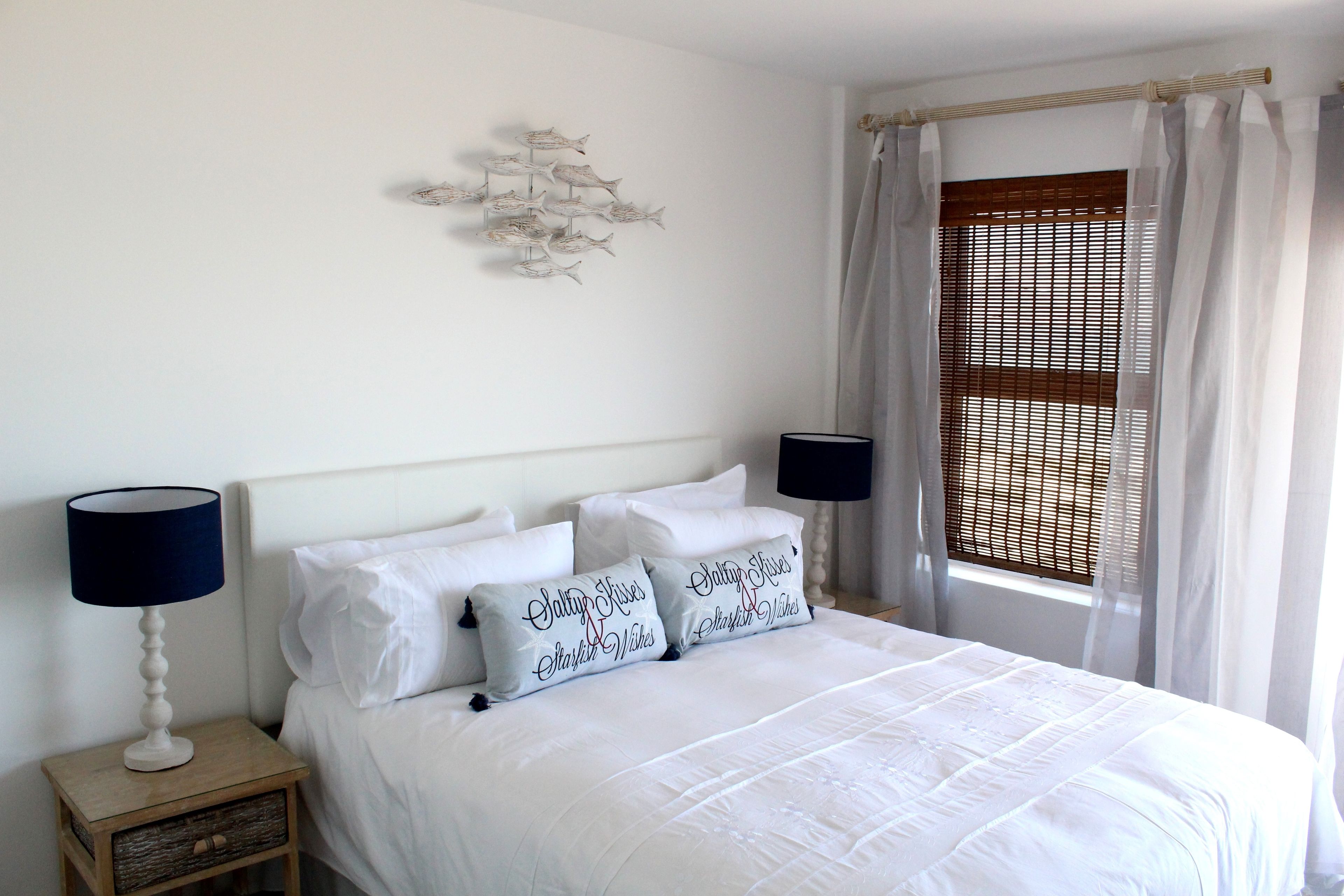 Una cama bien hecha con ropa de cama blanca y almohadones decorativos, una mesita de noche con lámpara y un adorno de peces en la pared.