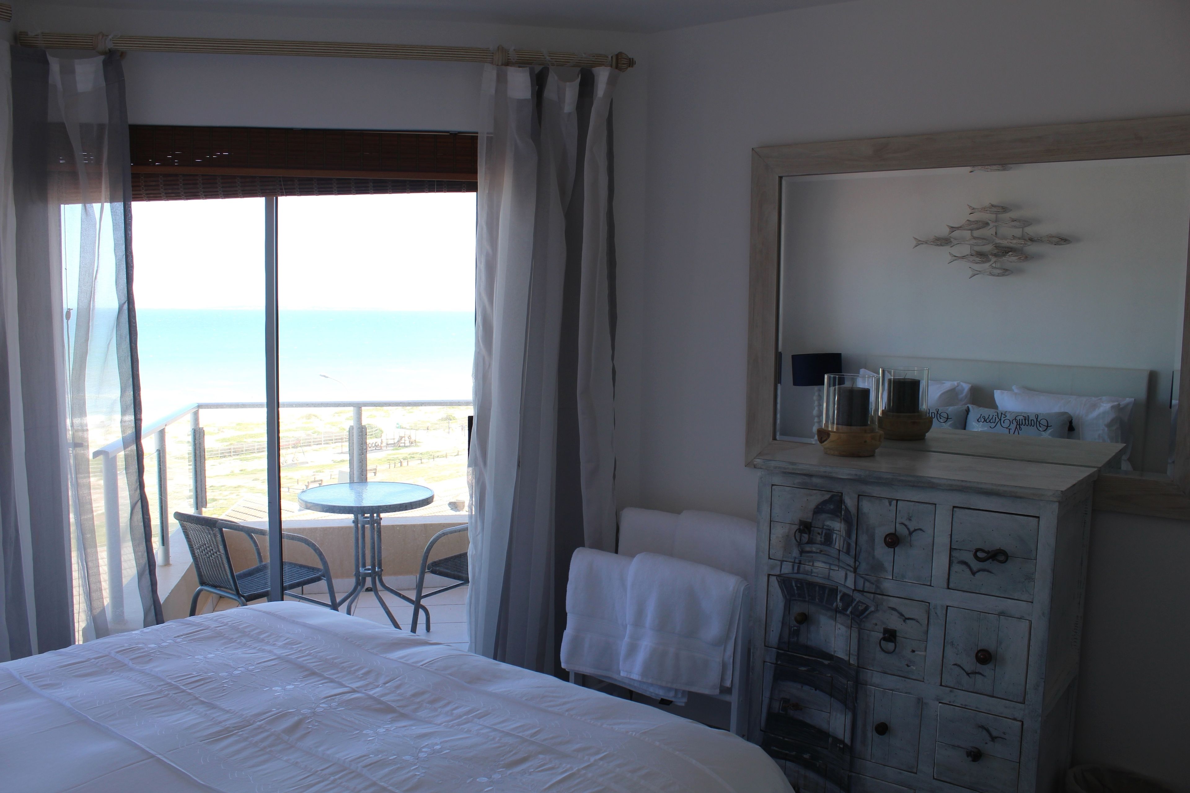 A hotel room with a bed, a small round table, a chair, and a view of the sea through large windows.