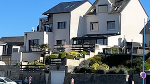 A modern two-story house with a flat roof, large windows, and a stone wall in front.