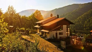 Un bâtiment de style chalet, doté d’un toit en pente, entouré d’une végétation luxuriante et avec des collines en arrière-plan.
