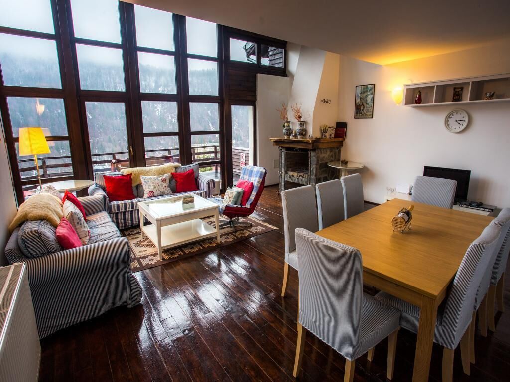 A cozy living room with a sofa set, a coffee table, and a dining table with chairs. There is a fireplace and a large window with a view of the outdoors.