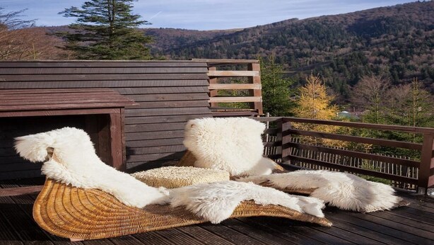 Une terrasse en bois avec un fauteuil de jardin en osier et de moelleux coussins blancs, donnant sur une colline boisée.