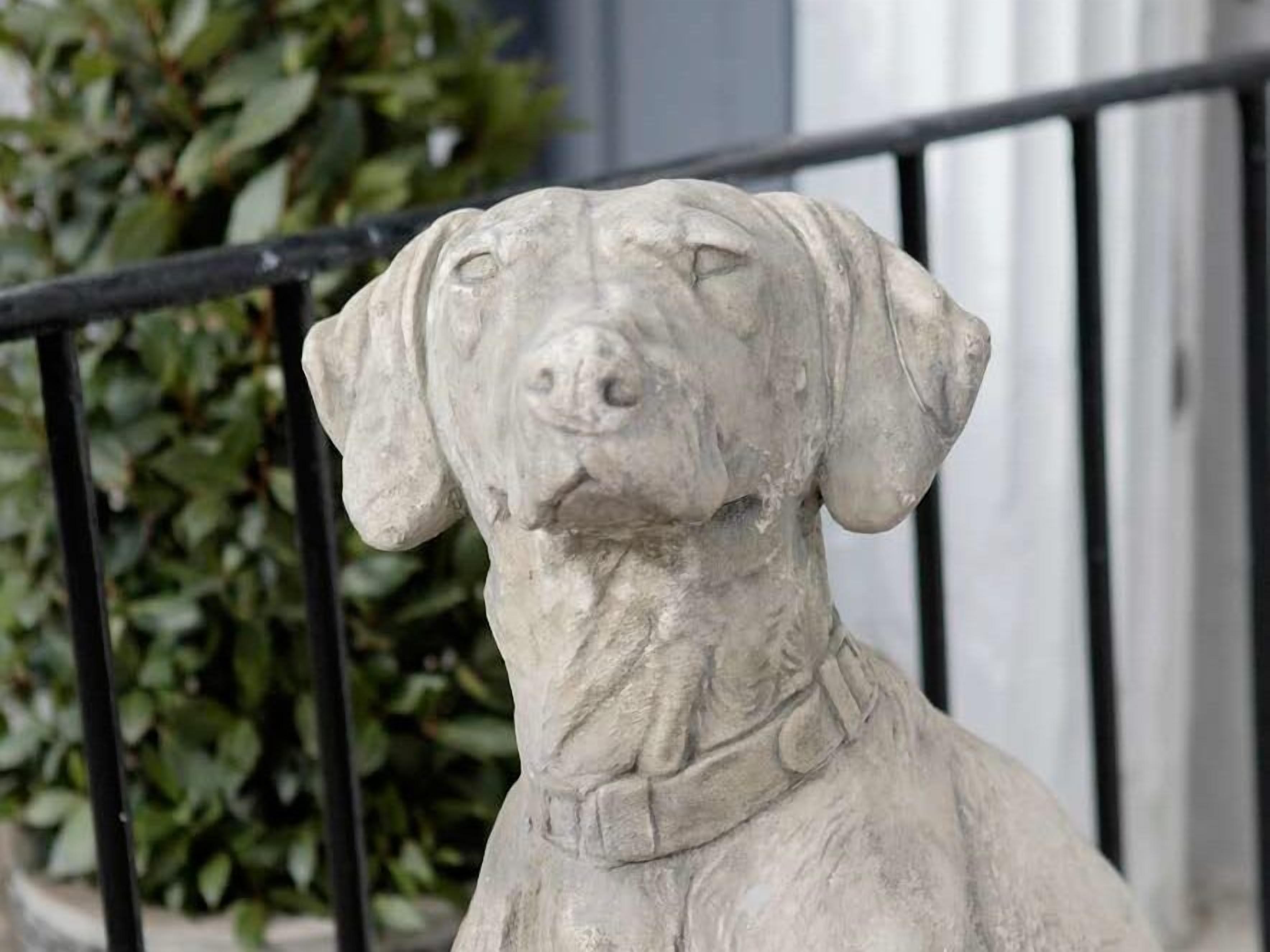Una estatua de piedra de un perro con collar, ubicada frente a una cerca de metal negro y un edificio de fachada blanca.