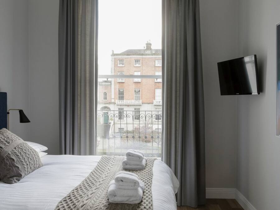 A hotel room with a bed, a TV mounted on the wall, and a view of a building through the window.