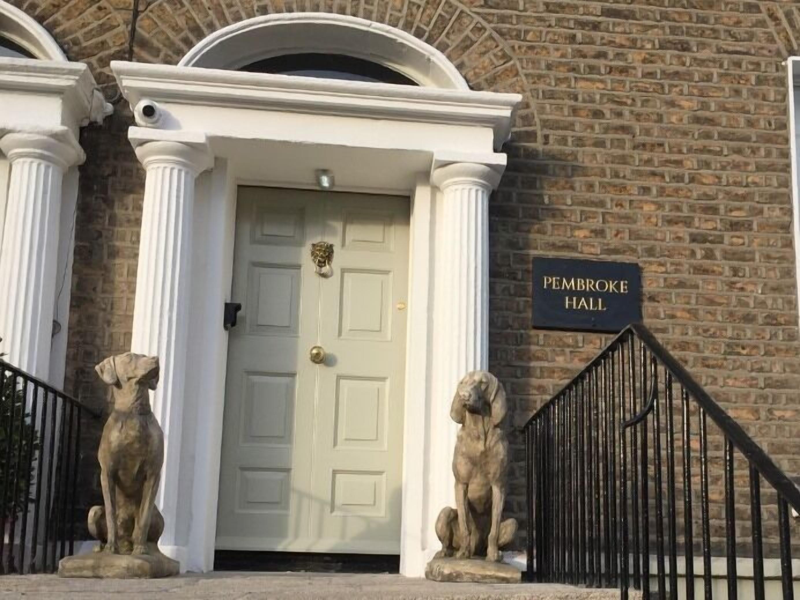 Una puerta blanca con un llamador de latón, flanqueada por dos estatuas de leones de piedra, y un cartel que dice "PEMBROKE HALL" sobre un muro de ladrillo.