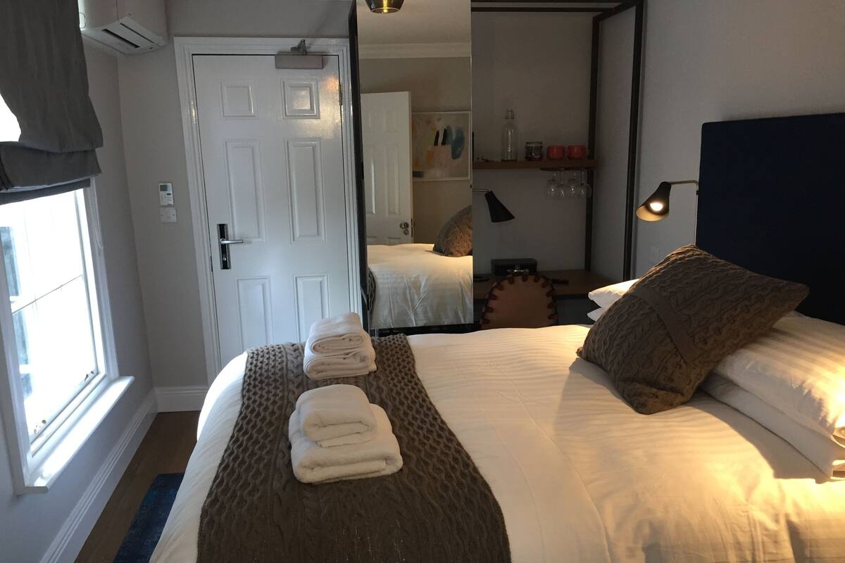 Una habitación de hotel con una cama grande, una mesita de noche, un espejo, una estantería con vasos y una ventana con cortinas.
