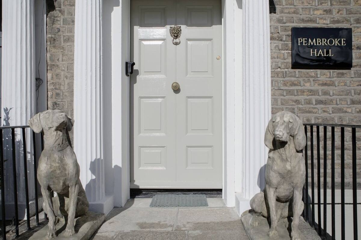 Una puerta blanca con un cartel que dice "Pembroke Hall" encima, flanqueada por dos estatuas de piedra con forma de perros, con un muro de ladrillo y una valla metálica al fondo.