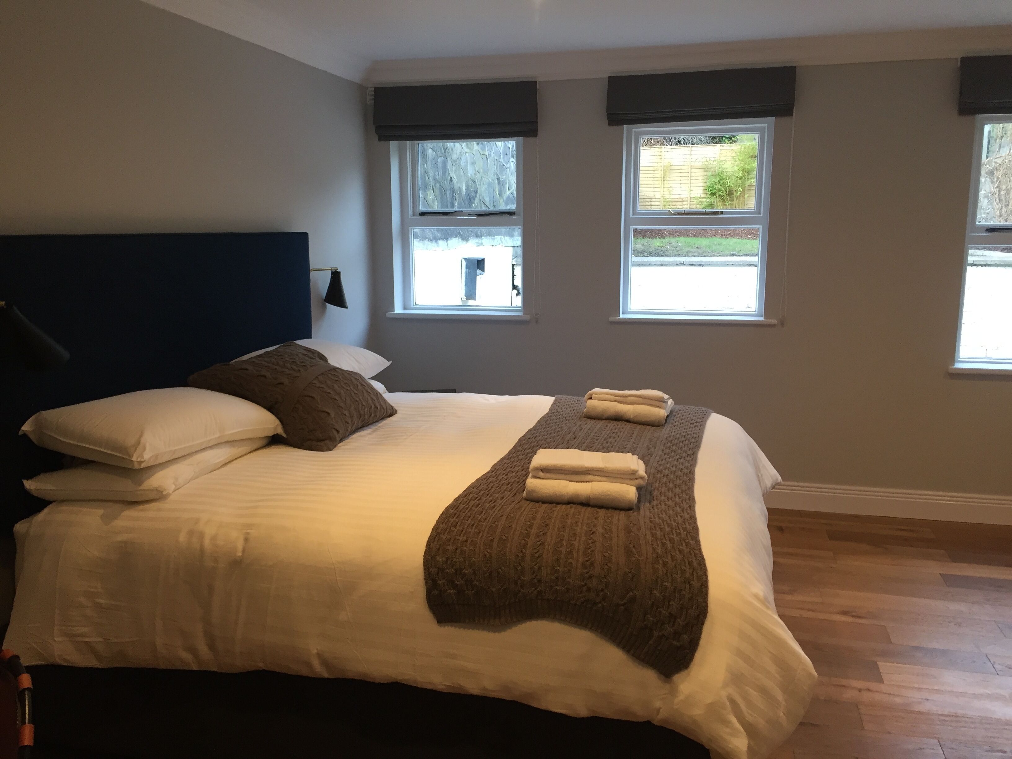 Una cama bien hecha con almohadas y una colcha, dos ventanas con persianas y una lámpara en la pared.