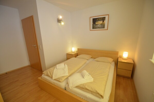 Un lit bien fait, avec deux oreillers et deux serviettes enroulées, une tête de lit en bois, deux tables de chevet avec des lampes, un tableau encadré au mur et une porte en bois.