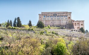 A large, historic building with multiple levels and arched windows, surrounded by a hilly landscape with sparse vegetation. - La Piaggia (Assisi)