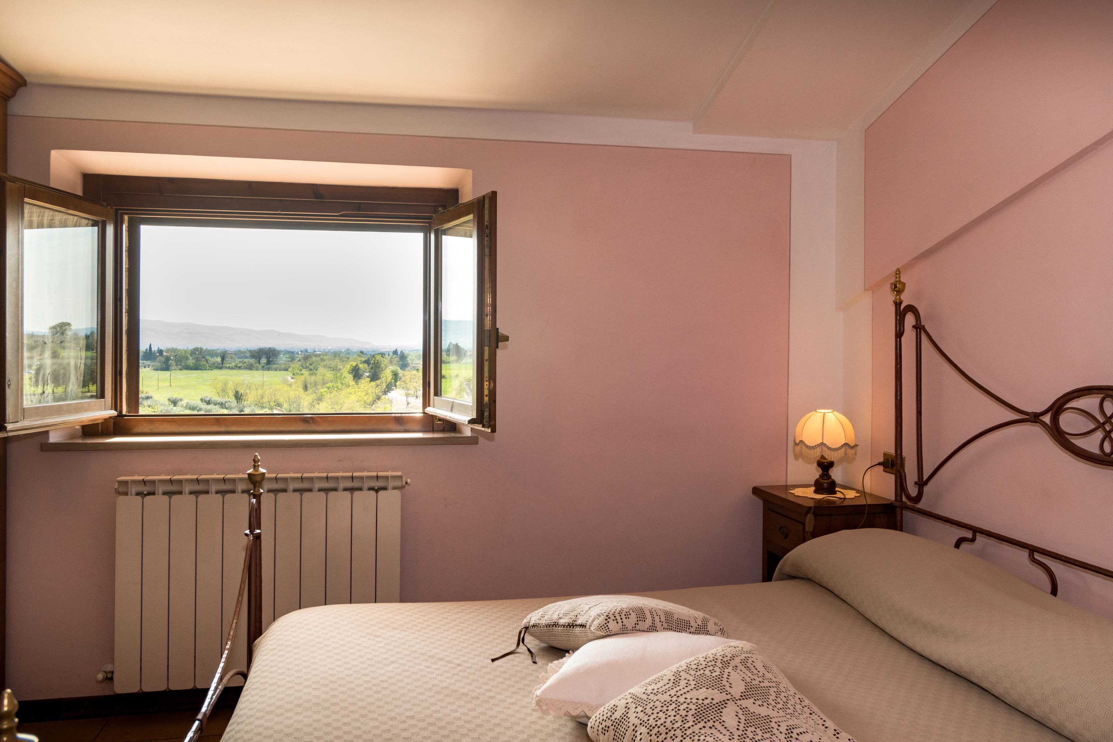 A bedroom with a large window, a radiator, a bedside table with a lamp, and a bed with pillows.