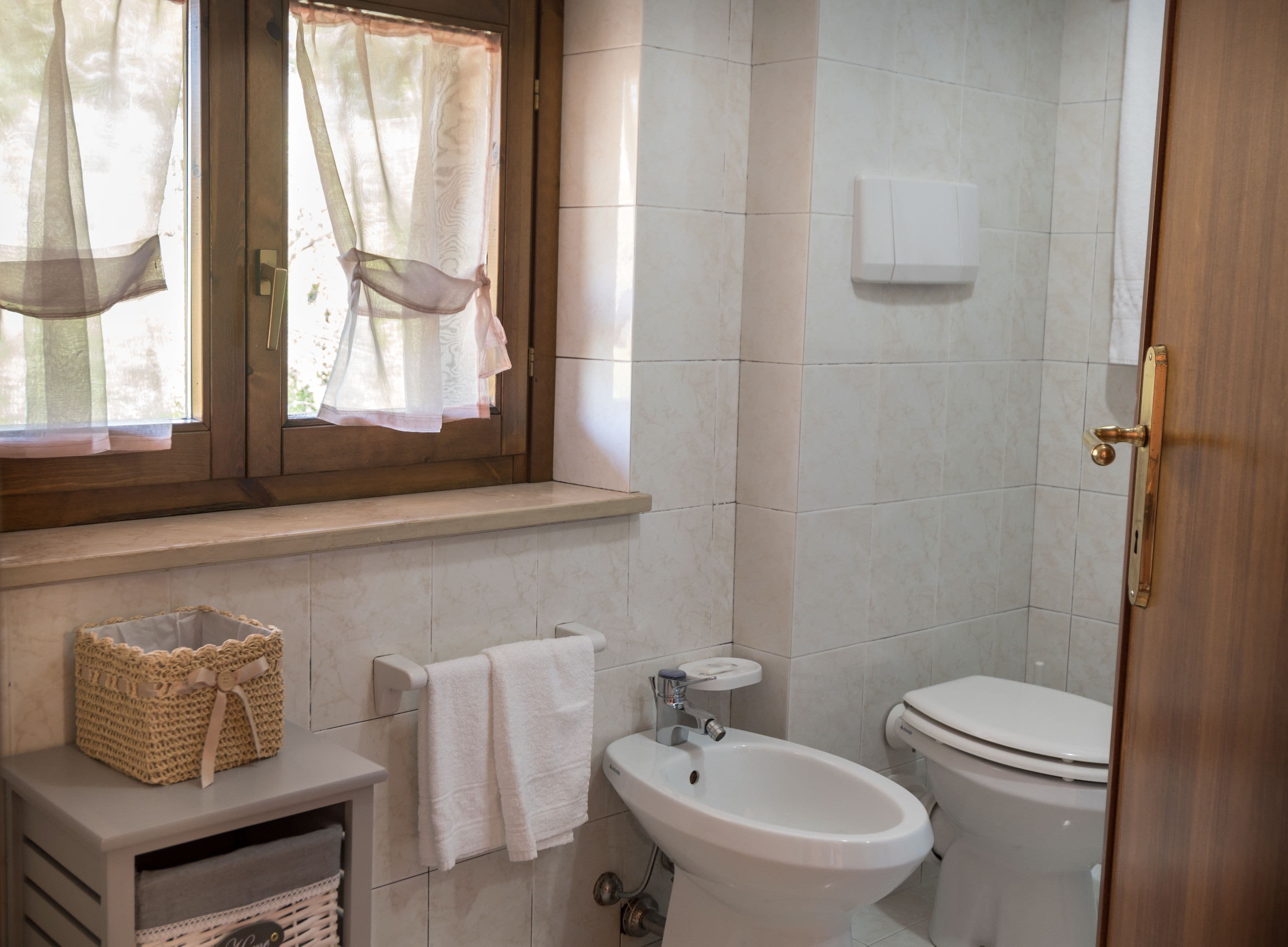 A bathroom with a toilet, bidet, sink, and a window with sheer curtains.
