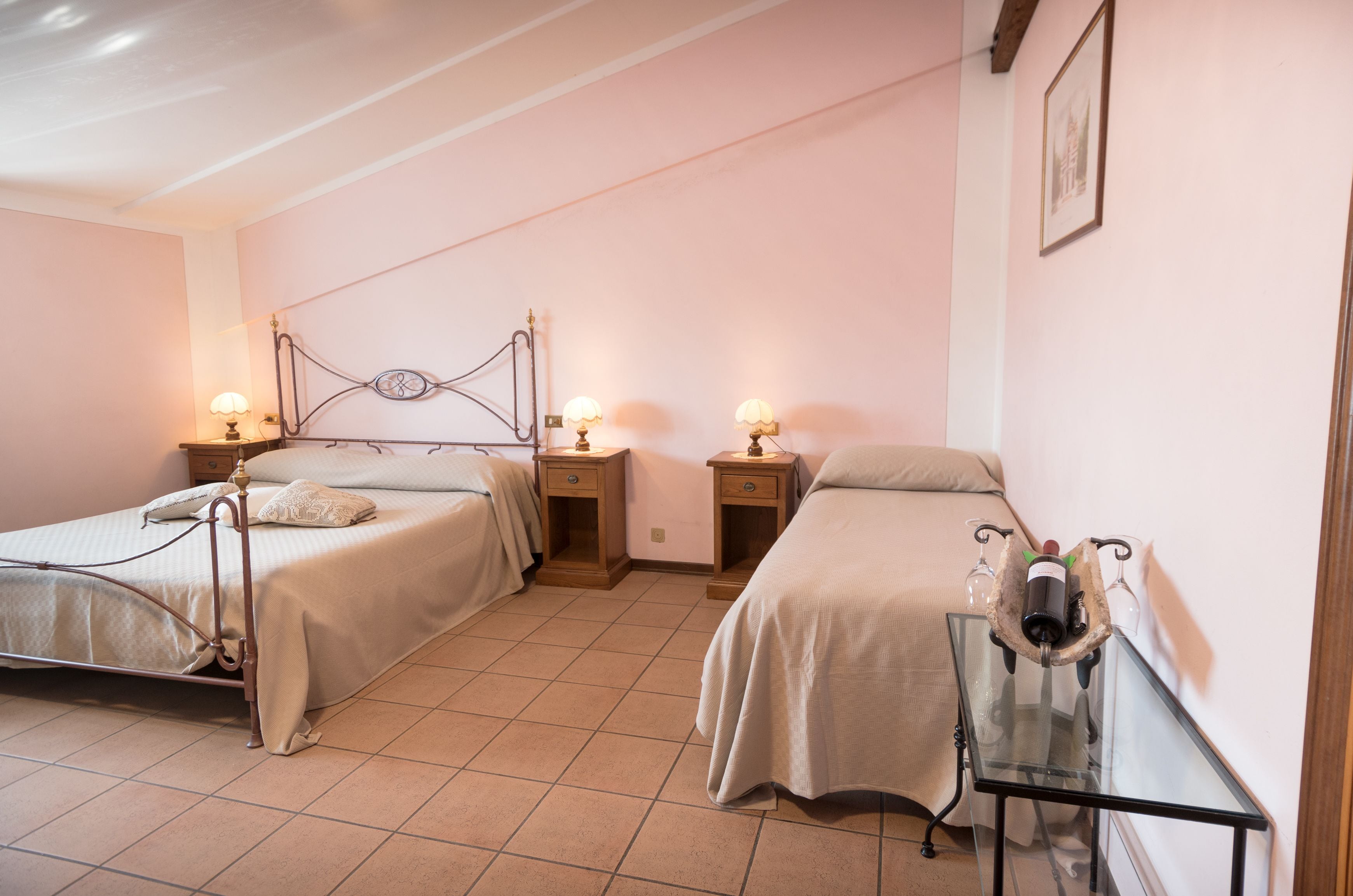A bedroom with two beds, bedside tables with lamps, a glass table with a wine bottle and glasses, and a framed picture on the wall.