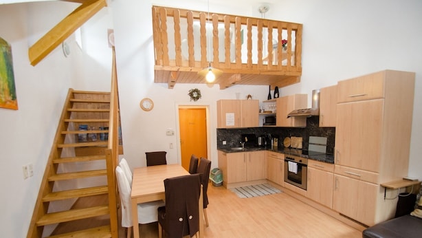 Een kleine keuken met houten kastjes, een eettafel met stoelen, een inbouwoven en een kleine trap naar een vide.