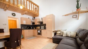 Een moderne keuken met houten kastjes, een inbouwoven en een magnetron. Er is een eettafel met stoelen en een bank in de woonkamer.