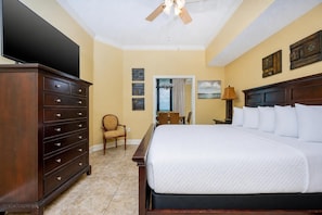 Quarto de hotel com uma cama grande, cômoda de madeira, uma cadeira, escrivaninha com luminária e obras de arte nas paredes.