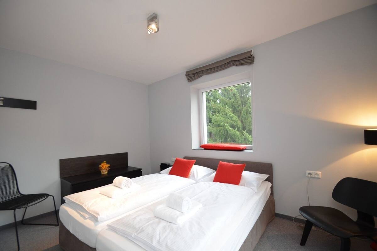 Habitación de hotel con una cama grande, una ventana con vistas a árboles, una mesita de noche y una silla.