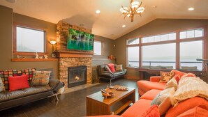 A spacious living room with a stone fireplace, a large flat-screen TV, leather sofas, and a view of the water through large windows.