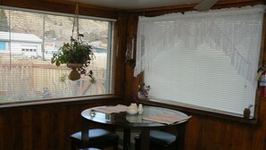 Een hoekkamer met een ronde tafel en vier krukken, houten wanden, een raam met zonwering en een hangplant.
