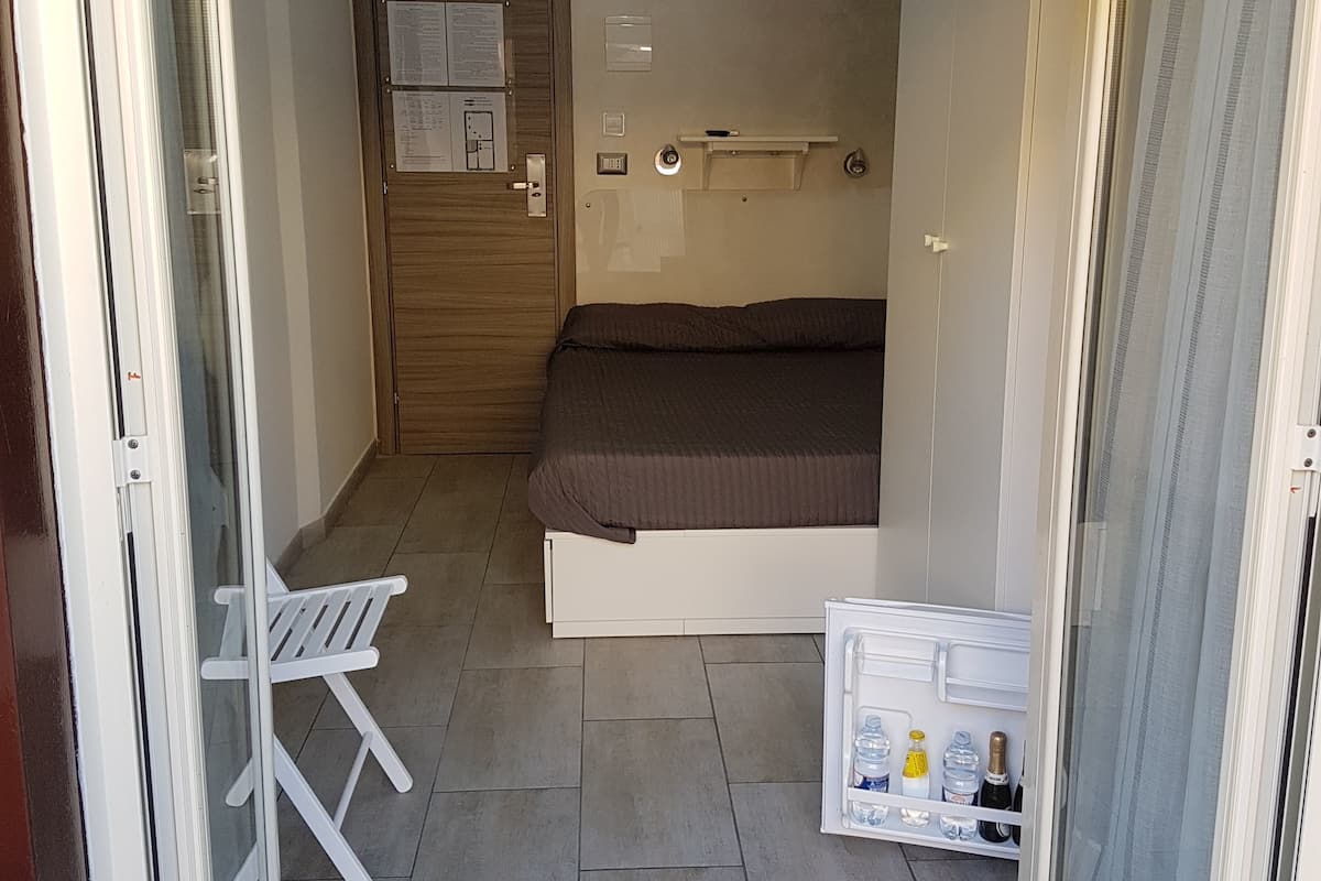 Habitació petita amb un llit individual, una porta de fusta, una tauleta de nit blanca i una petita estant amb ampolles, i una cadira blanca.