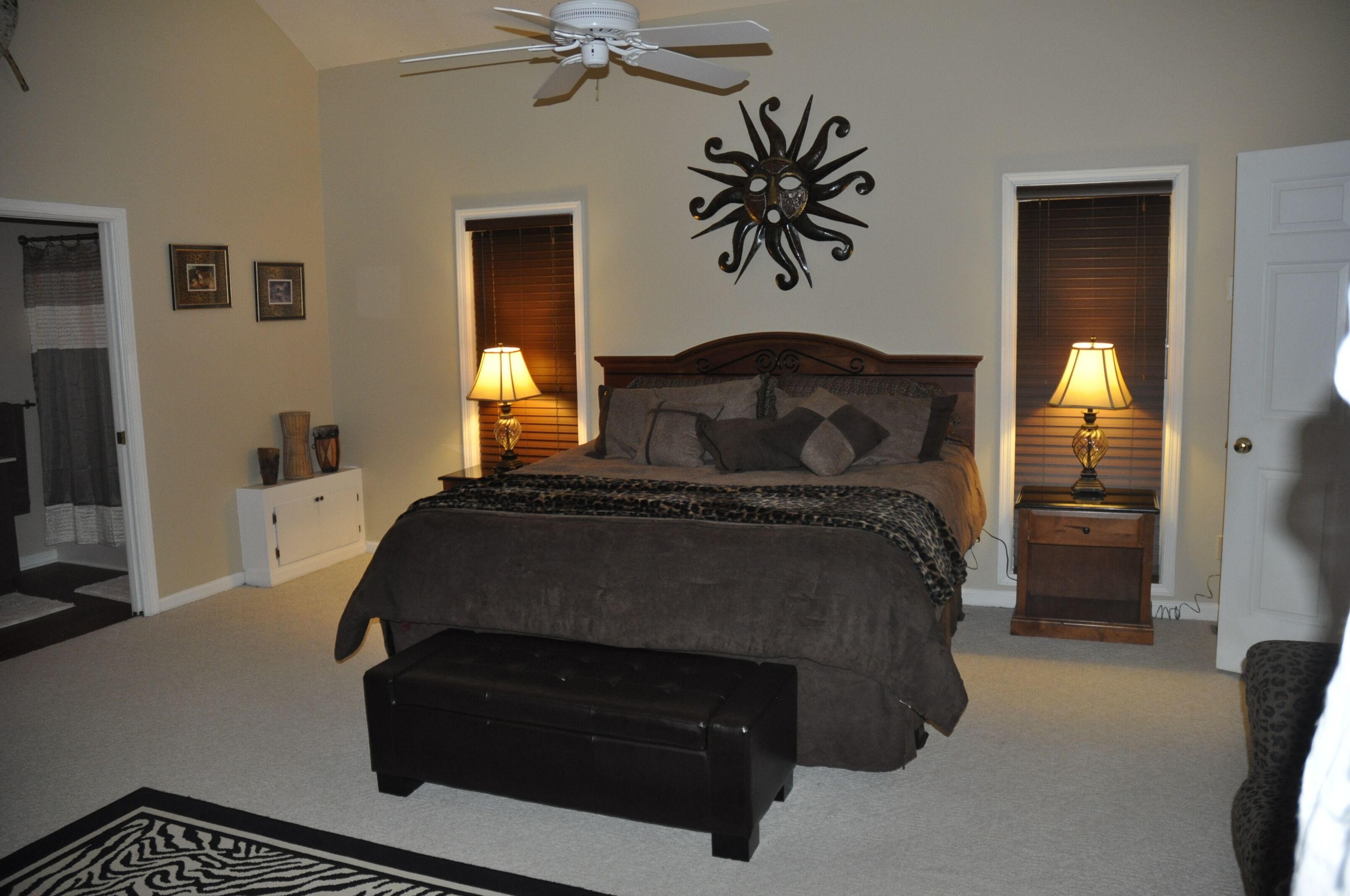 A bedroom with a bed, two bedside lamps, a ceiling fan, a decorative wall piece, a bench, and a door.