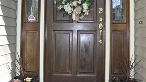 Una puerta de madera con un adorno de guirnalda, dos macetas con plantas y un felpudo.