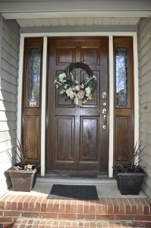A wooden front door with a wreath, two potted plants, and a doormat.